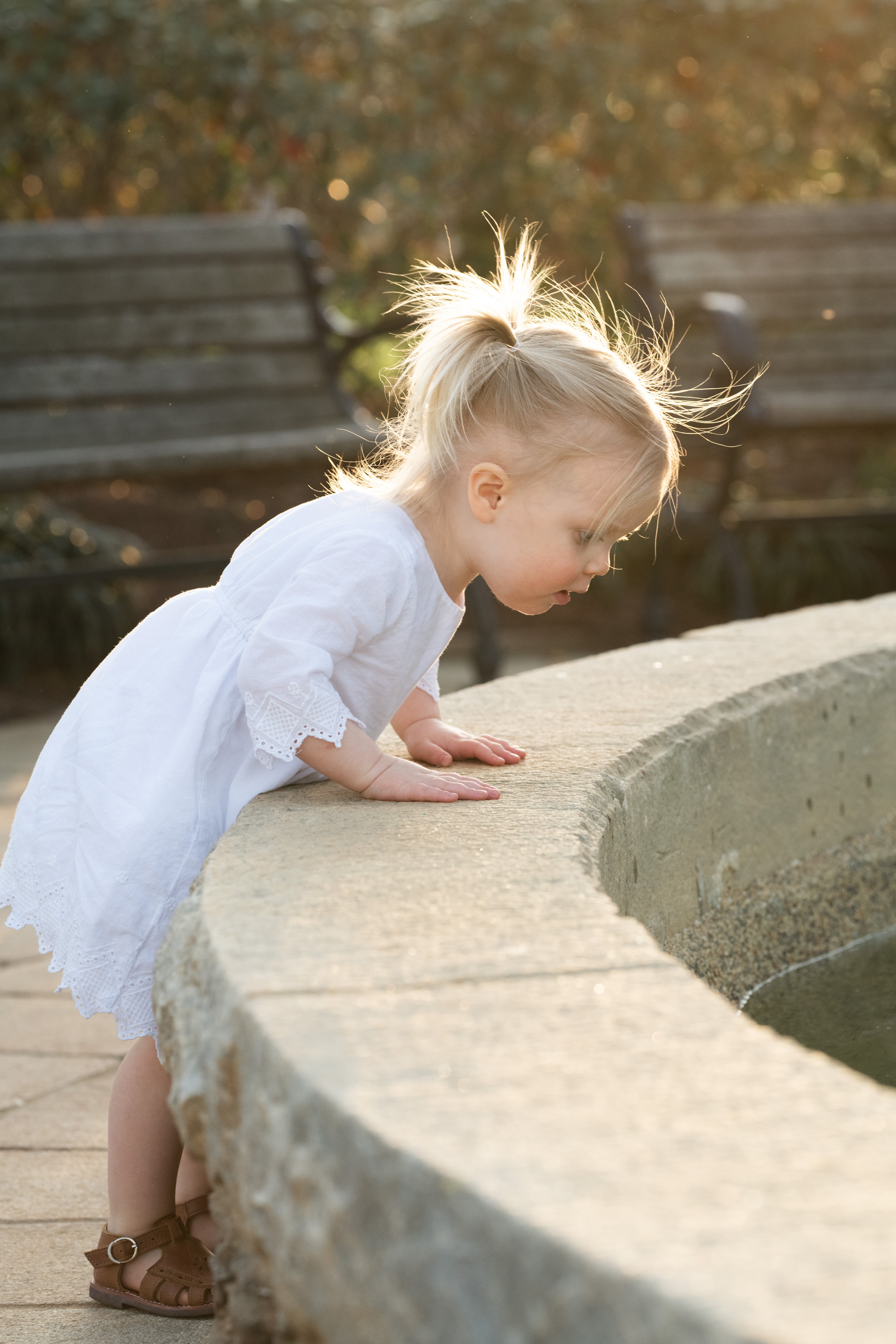 Toddler girl looking into a fountain at a park at sunset. The girl has blonde hair and is wearing a white dress with brown sandles.