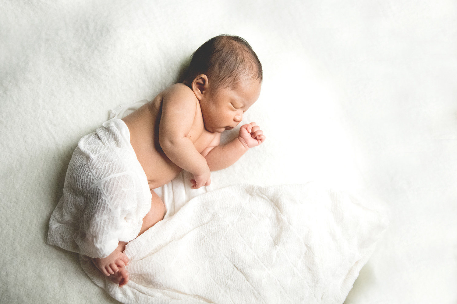 Asian newborn baby partially covered in an off-white knit blanket lying on her side on a white blanket.