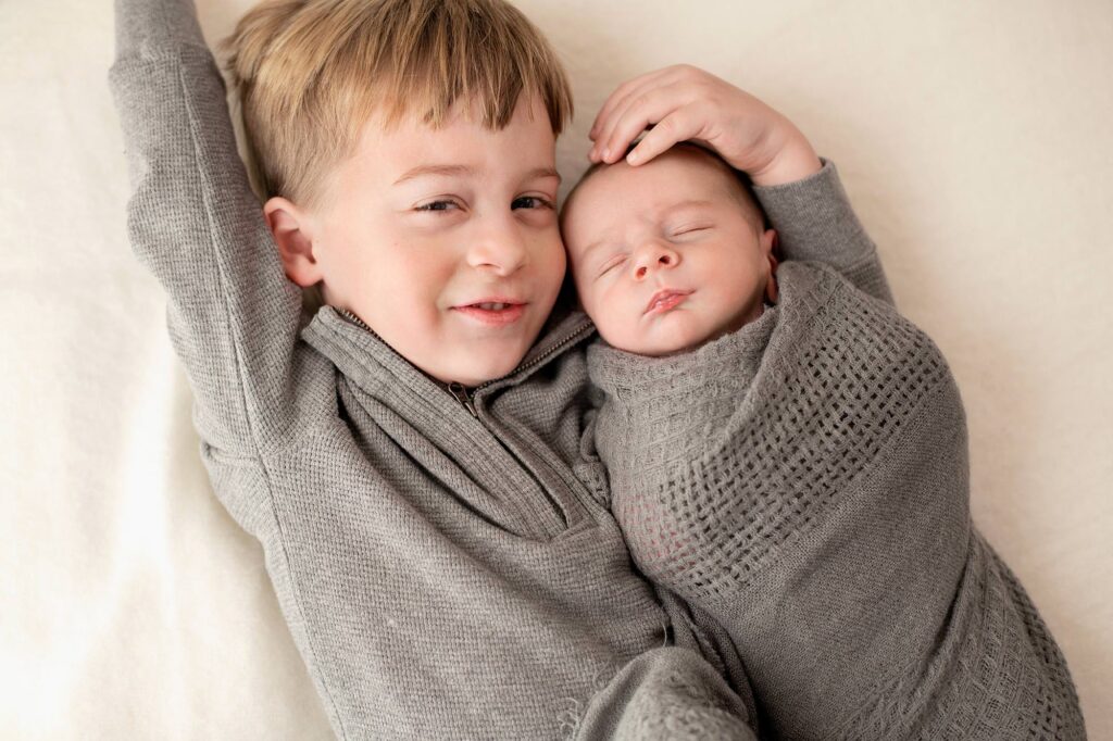 How to prepare for your Newborn Photo Session, Sibling brothers, one wearing a grey quarter zip and the newborn wrapped in a grey swaddle. 